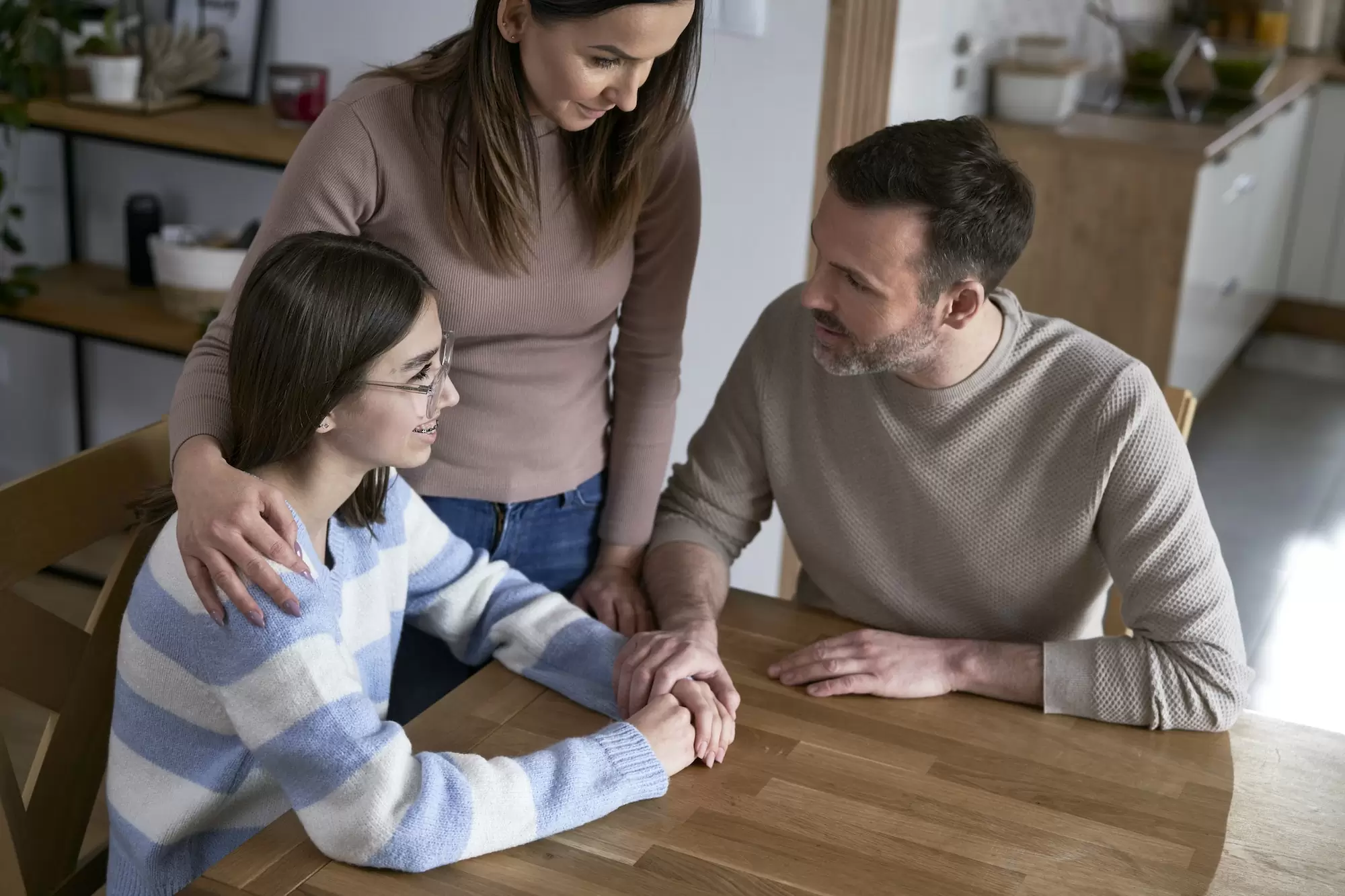 Two parents sitting at a kitchen table, gently talking with their teenage daughter